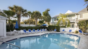 A swimming pool with lounge chairs and umbrellas, surrounded by palm trees and a white picket fence.