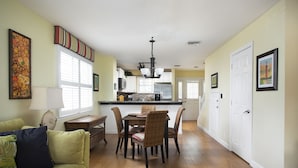 A spacious living room with a green sofa, a dining table with chairs, and a kitchen area in the background.