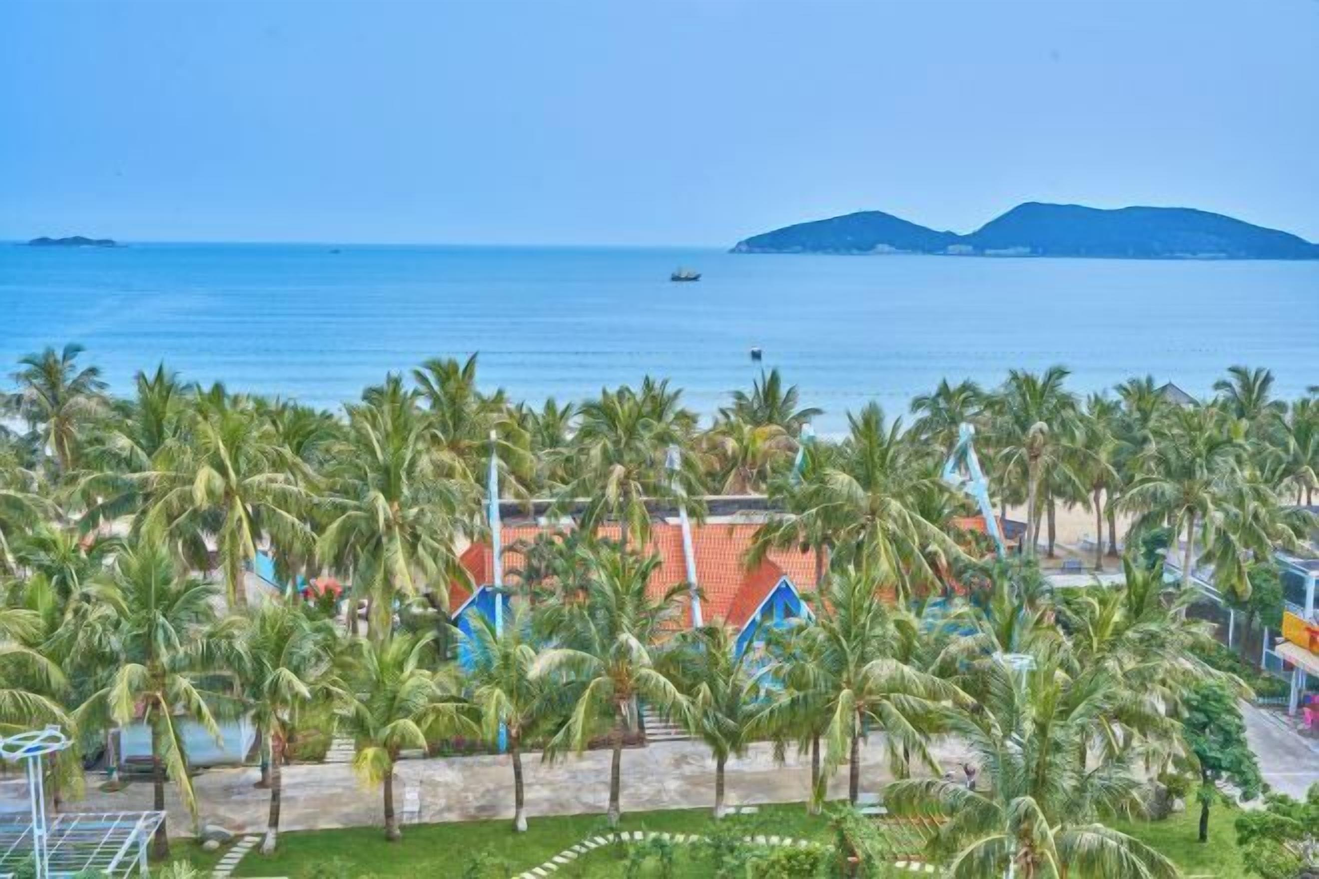 A coastal view with palm trees, a beach, and a distant mountain range.