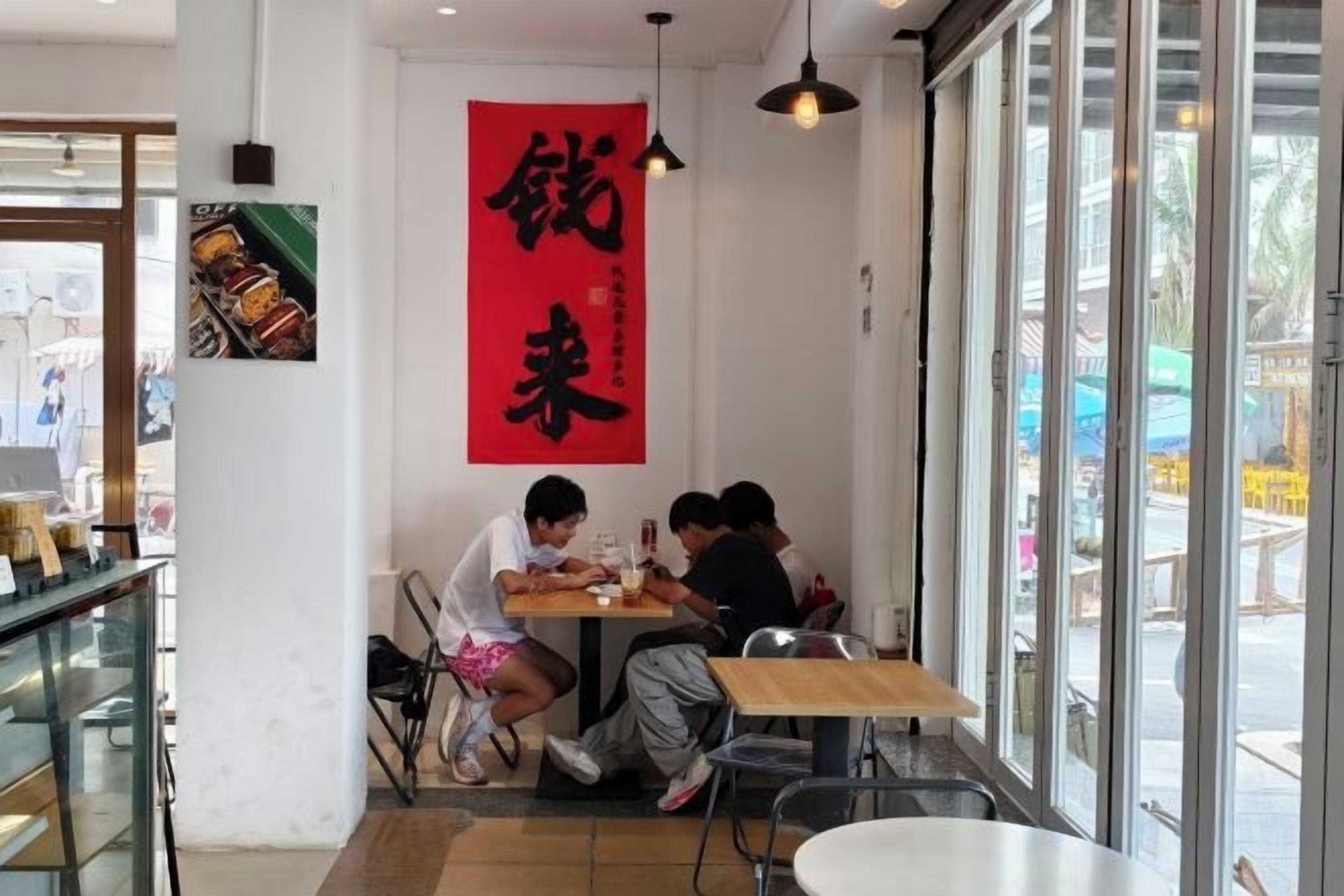 Ein Restaurantinneres mit einer Gruppe von Personen, die an einem Tisch essen, einem großen roten Banner an der Wand und Blick durch das Fenster.
