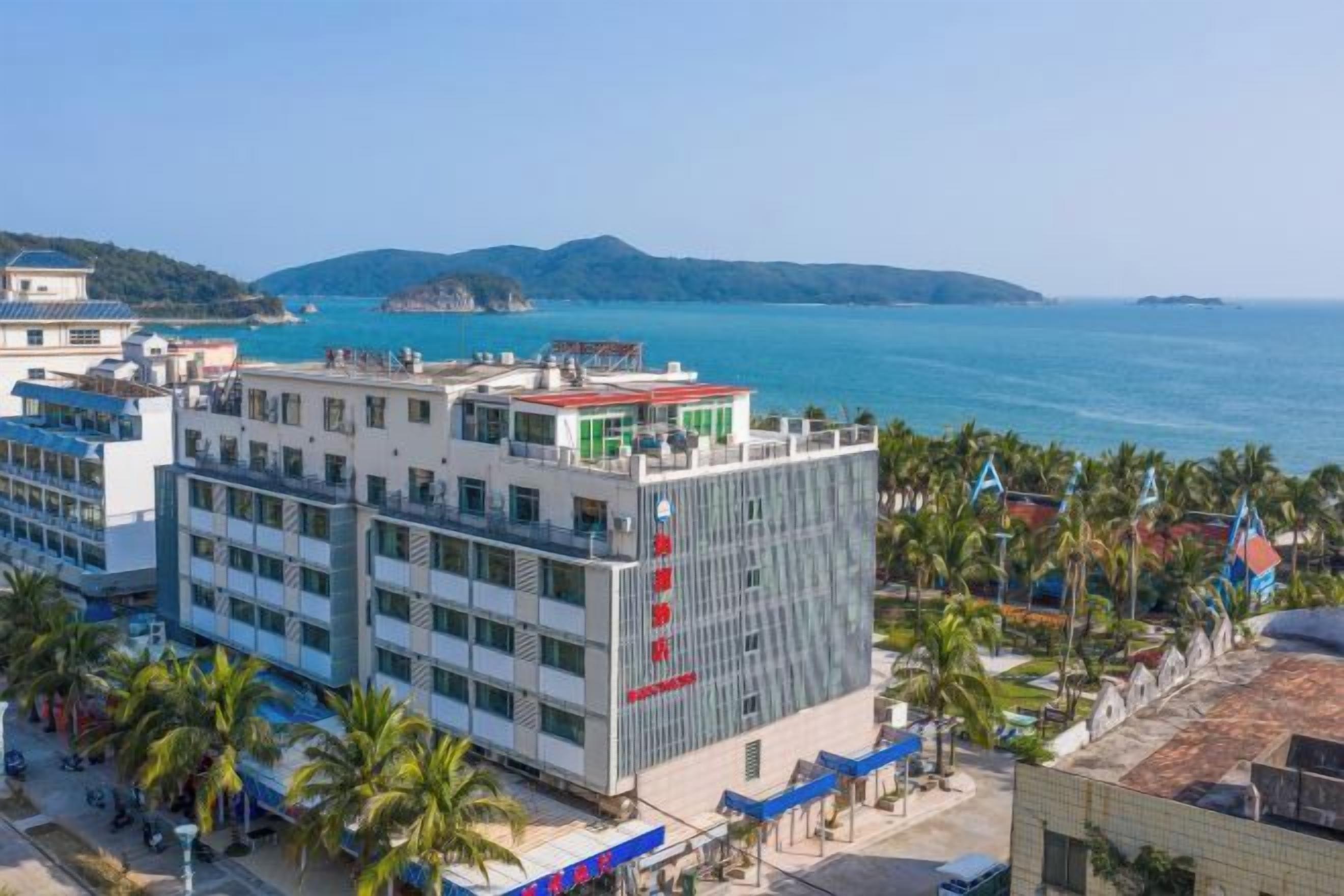 A multi-story hotel with a view of the sea, surrounded by palm trees and a hilly landscape.