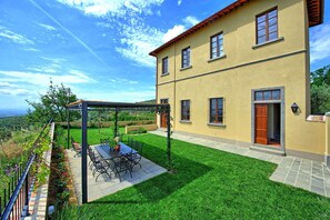 Ein zweigeschossiges Gebäude mit einer Terrassenfläche, die einen Tisch und Stühle beinhaltet, umgeben von einem gepflegten Rasen und einer Pergola.