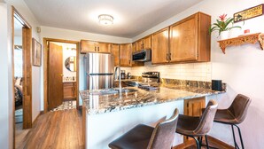 A kitchen with wooden cabinets, a granite countertop, a stainless steel refrigerator, and a microwave. There are two bar stools and a potted plant on the countertop.