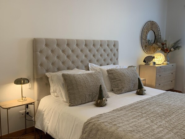 A neatly made bed with a tufted headboard, a bedside lamp, and a small nightstand with a decorative lamp and vase.