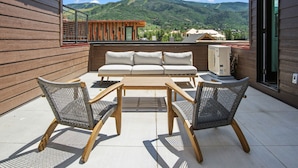 A patio with a sofa, two chairs, and a table. A large air conditioning unit is visible. - Urban Luxury - Rooftop Patio w/ Hot Tub, Perfect for 2 Families at Competitive Rates (Steamboat Springs)