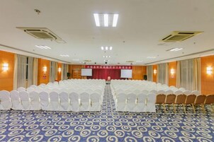 Un salón de conferencias con filas de sillas, un escenario con cortina roja y una alfombra de estampado azul.