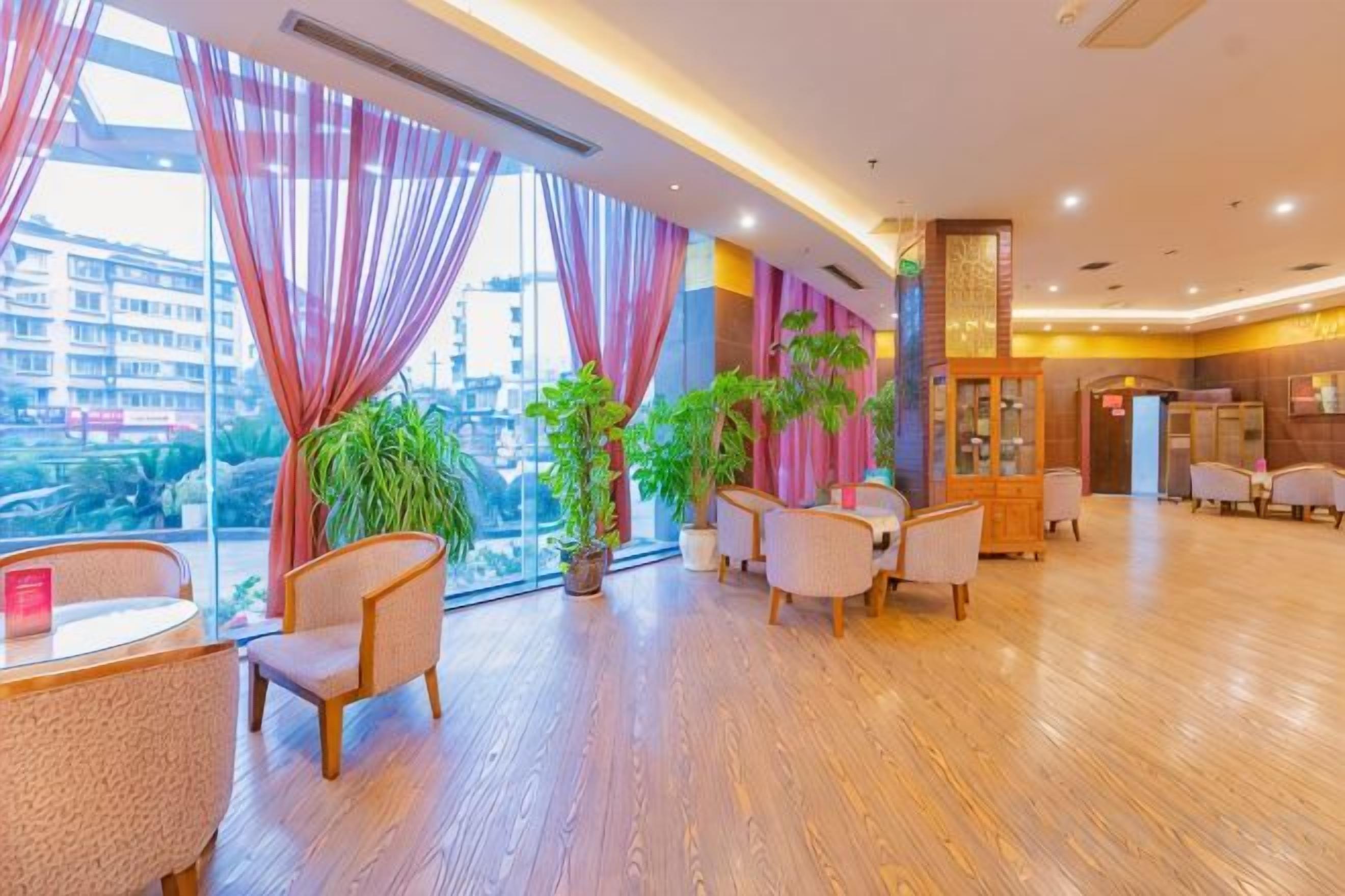 A hotel lobby with a wooden floor, large windows with curtains, and several potted plants.