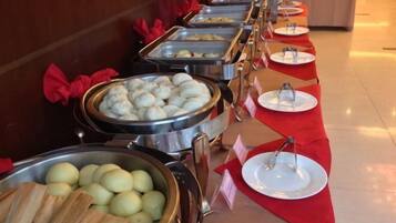 Una barra de buffet con distintos alimentos en recipientes metálicos, sobre un mantel rojo con platos y cubiertos de plata.