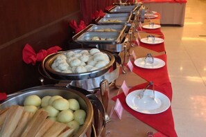 Una barra de buffet con distintos alimentos en recipientes metálicos, sobre un mantel rojo con platos y cubiertos de plata.