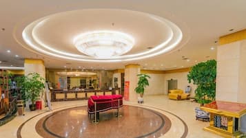 A hotel lobby with a central circular seating area, a reception desk, and potted plants.