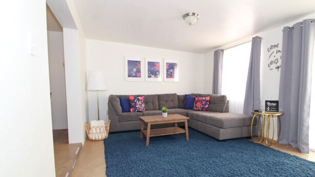 Una sala de estar moderna con un sofá seccional, una mesita de madera y un tapete azul. Hay tres cuadros enmarcados en la pared, una lámpara y una mesita auxiliar con un letrero.