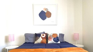 Un dormitorio con colcha azul, dos almohadas con diseños abstractos, una obra de arte enmarcada sobre la cama y dos lámparas de noche.