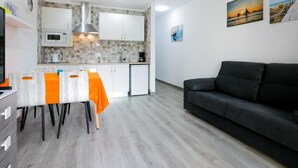 Una cocina moderna con armarios blancos, microondas y cafetera. Hay una mesa de comedor con sillas anaranjadas. Un sofá negro está en la zona de estar, y dos cuadros decoran la pared.