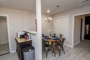 Ett kompakt kök med mikrovågsugn, kaffebryggare och papperskorg. Ett matbord med stolar och en vas med blommor står i mitten.
