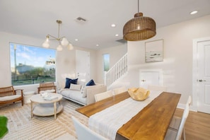 Uma sala de estar moderna com uma mesa de jantar de madeira, sofás brancos, cadeiras de vime e uma grande janela com vista para o exterior.