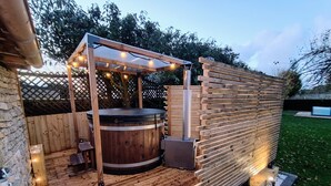 Un área de hidromasaje exterior de madera con una pérgola en la parte superior, rodeada por un cerco de madera y un muro de piedra.