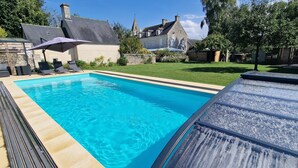 Una piscina con sillones de descanso y una zona cubierta, rodeada por un césped cuidado y edificios de piedra.