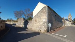 A stone building with a sloped roof, a street in front, and a church in the background.