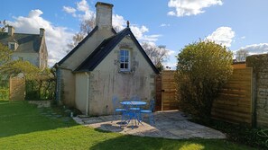 A small house with a stone chimney, a window, and a small door. There is a paved patio with a table and chairs.
