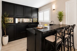 A kitchen with black cabinetry, a central island with bar stools, a potted plant, and a window with blinds.