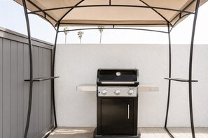 Ein Grill mit Sonnendach vor einer schlichten Wand, an deren Seite ein Zaun steht.