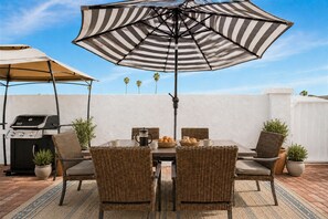 An outdoor patio with a grill, wicker chairs, a striped umbrella, and a white wall backdrop.