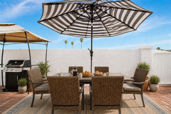 An outdoor patio with a grill, wicker chairs, a striped umbrella, and a white wall backdrop.