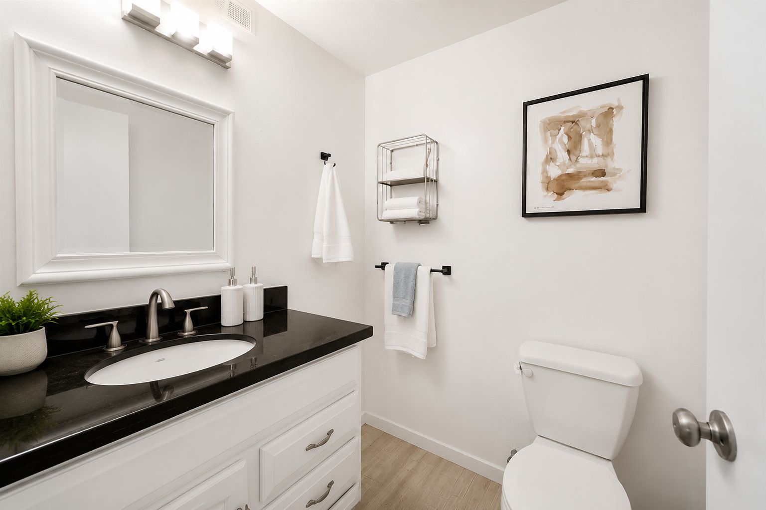 A modern bathroom with a black countertop sink, white cabinets, a toilet, and a framed abstract painting on the wall.