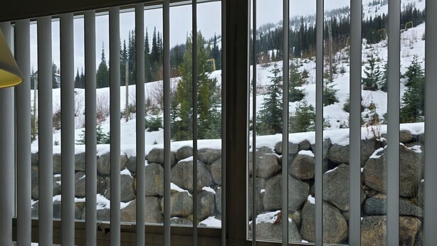 A view through a window with vertical blinds, featuring a snowy landscape with trees and a stone wall. - Apex Inn 223 sleeps 5. Ski in/Ski out. Steps to Apex Resort Village (Penticton)