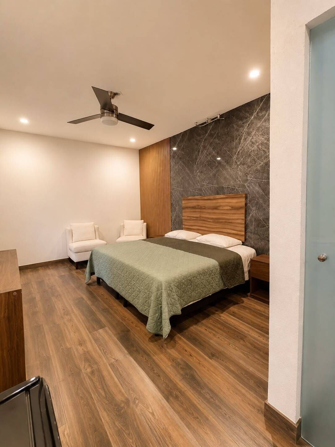 A modern bedroom with a bed, two chairs, a wooden floor, a ceiling fan, and a marble accent wall.
