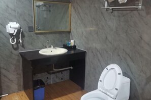 A bathroom with a marble wall, a white sink, a mirror, a toilet, and a trash bin. - Huilong Hotel (Sihui)