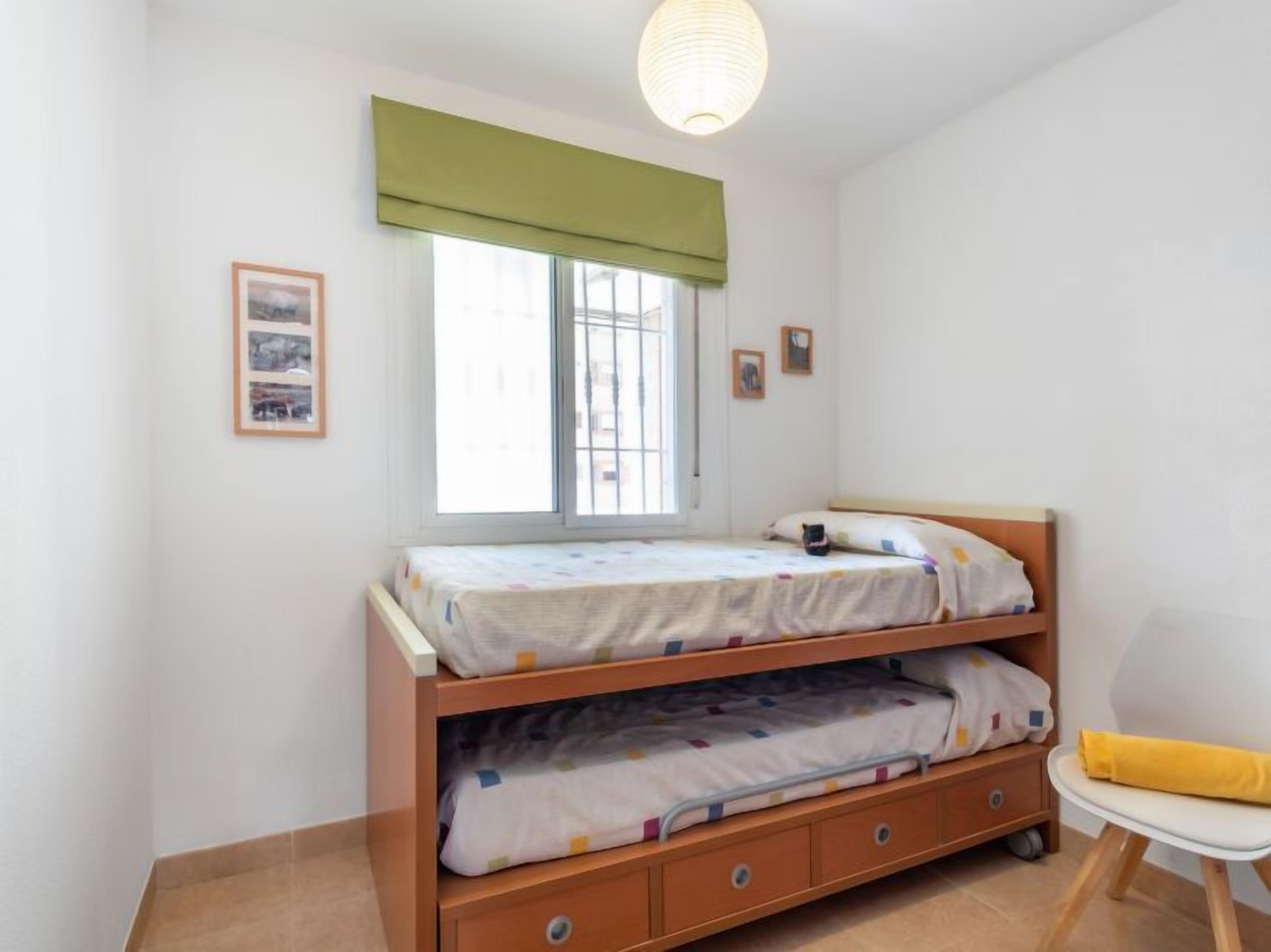 Een kleine, schone kamer met een stapelbed, een stoel en een raam met een groene zonwering.