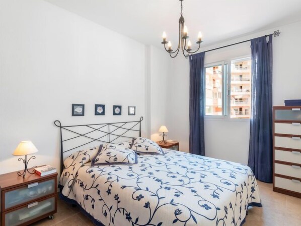 Un dormitorio con cama, mesitas de noche, cómoda, candelabro y ventana con cortinas.