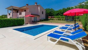 Uma casa moderna de dois andares com piscina, cadeiras de descanso azuis e guarda-sóis vermelhos.