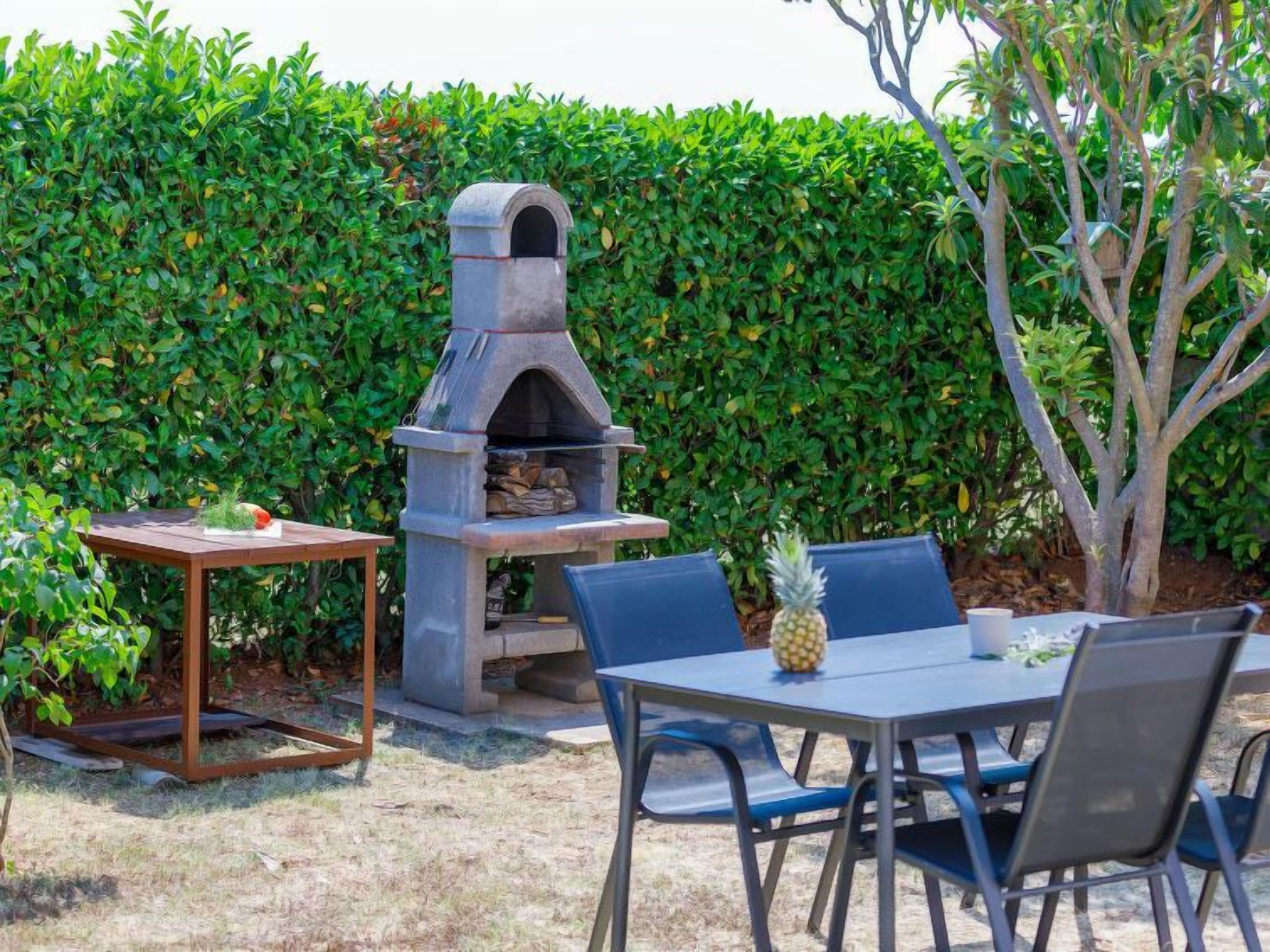An outdoor patio with a stone-built pizza oven, a table with chairs, and a neatly trimmed hedge in the background.