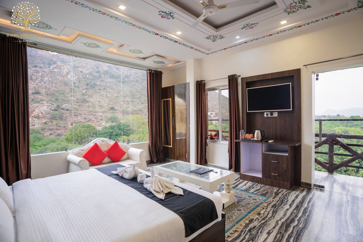 A hotel room with a large bed, a glass coffee table, a TV, and a view of the mountains.