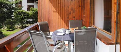 A balcony with a table set for two, chairs, and a window overlooking a garden.