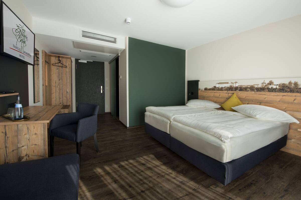 A hotel room with a bed, a desk, and a chair. There is a wardrobe and a door leading to another room.