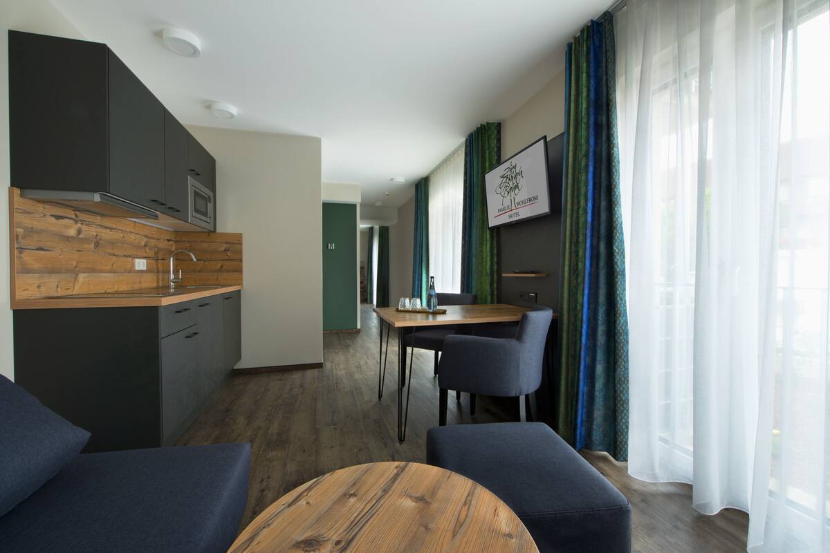 A modern hotel room with a kitchenette, a dining area, and a seating area with a wooden table.