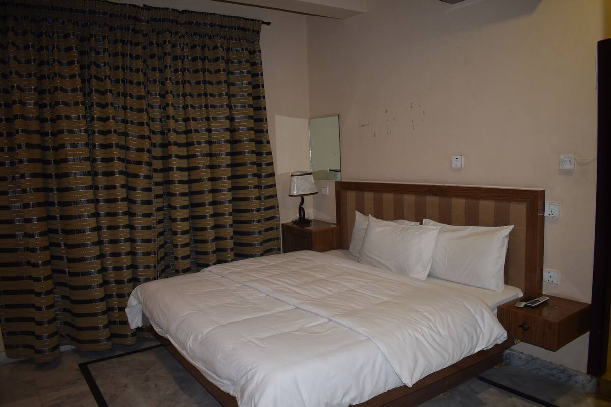 Una cama bien hecha en una habitación de hotel, con cabecera de madera, una mesita de noche con lámpara y un ventanal con cortinas.