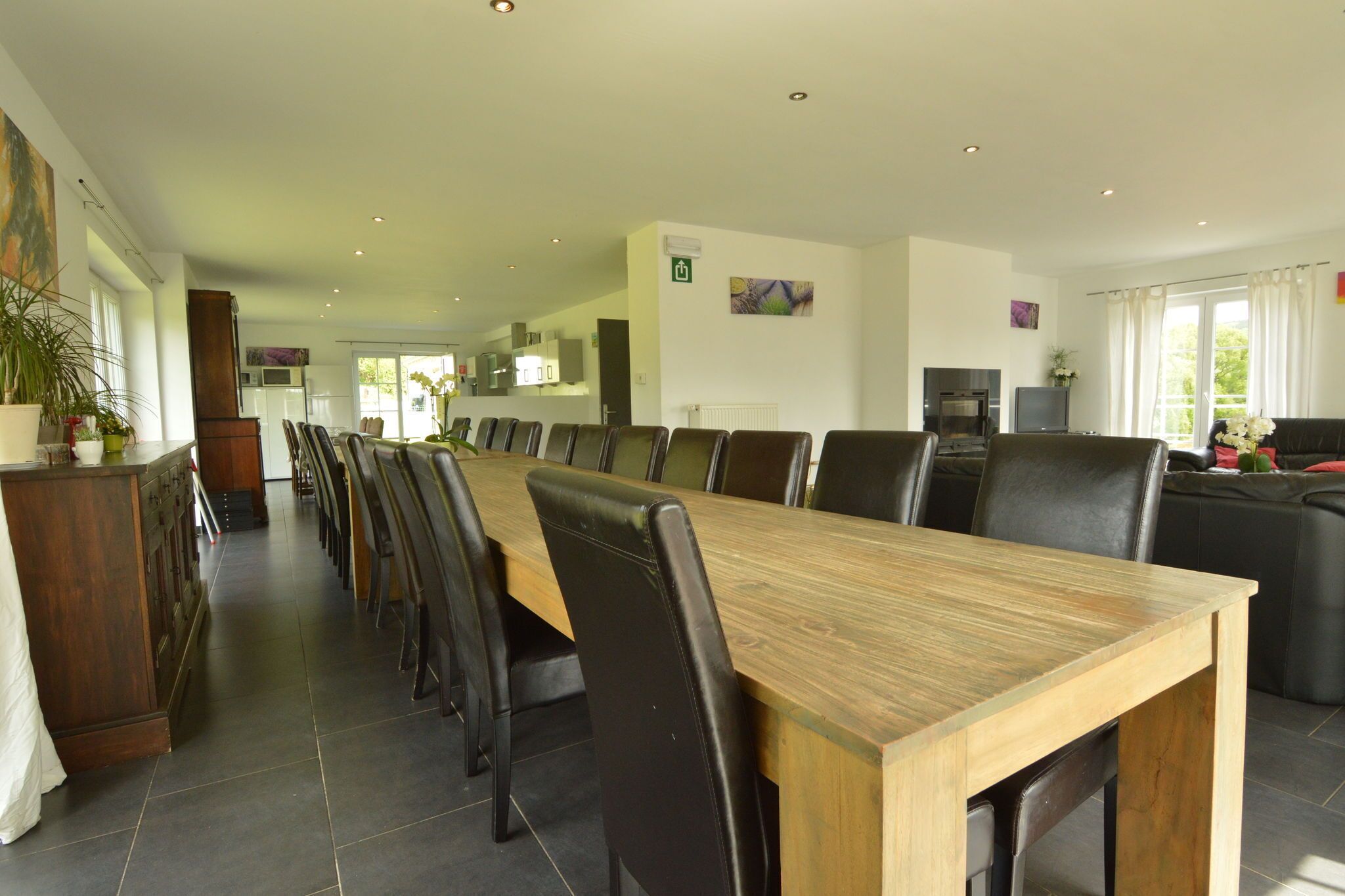 A spacious dining area with a long wooden table and black chairs, a kitchen in the background, and a living area with a sofa and a TV.