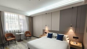 Habitación de hotel con una cama grande, dos mesitas de noche, una mesita redonda pequeña, dos sillas y un ventanal con cortinas.