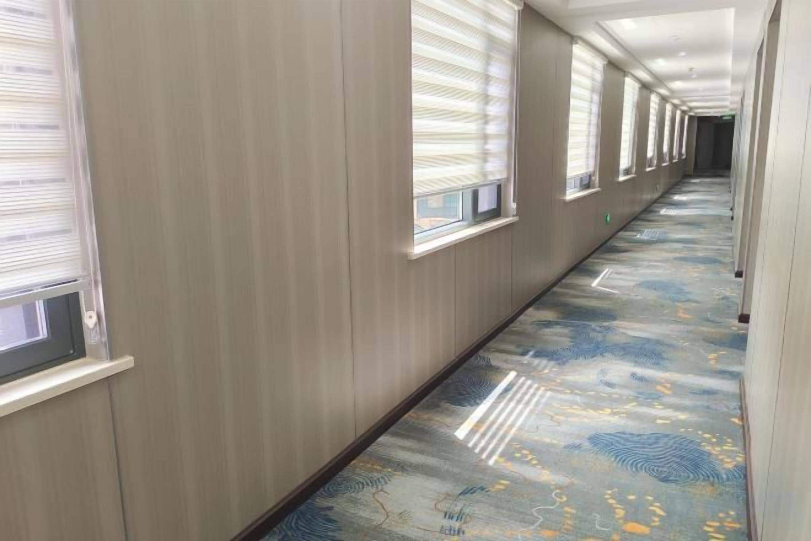 A hallway with a patterned carpet, wooden walls, and windows with blinds.