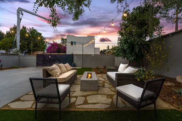 Une terrasse extérieure aménagée avec des meubles en osier, un foyer et des guirlandes lumineuses, le tout sur fond de coucher de soleil.