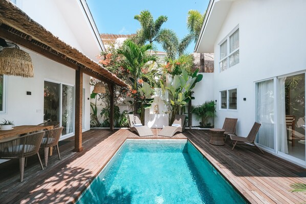 A pool area with a wooden deck, lounge chairs, and a thatched roof structure. - Sun & Moon Villa (Canggu)