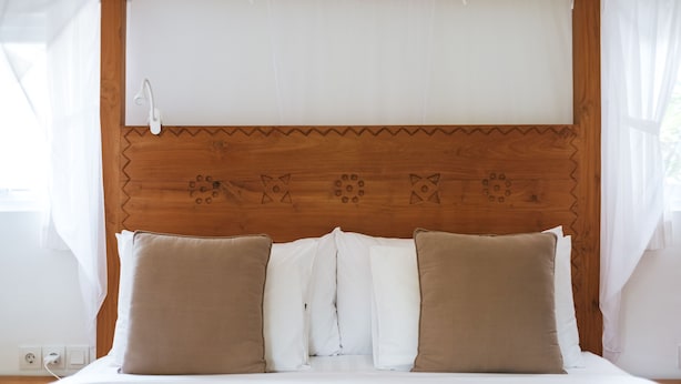 Una cama bien hecha con cabecera de madera con detalles decorativos, ropa de cama blanca y dos almohadas marrones.