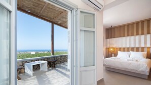 A bedroom with a bed, a bedside table, and a nightstand. There is a window with a view of the ocean and buildings. - New Villa Katerina Collection (Santorini)