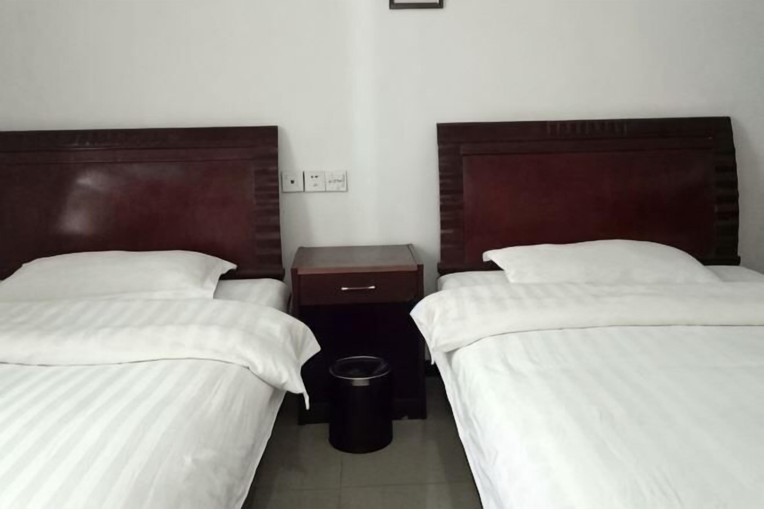A hotel room with two single beds, a nightstand, a trash bin, and a wall socket.