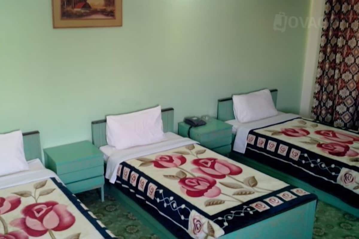 A room with three single beds, each with a floral bedspread and white pillows. There is a small table between the beds and a wall-mounted shelf above.