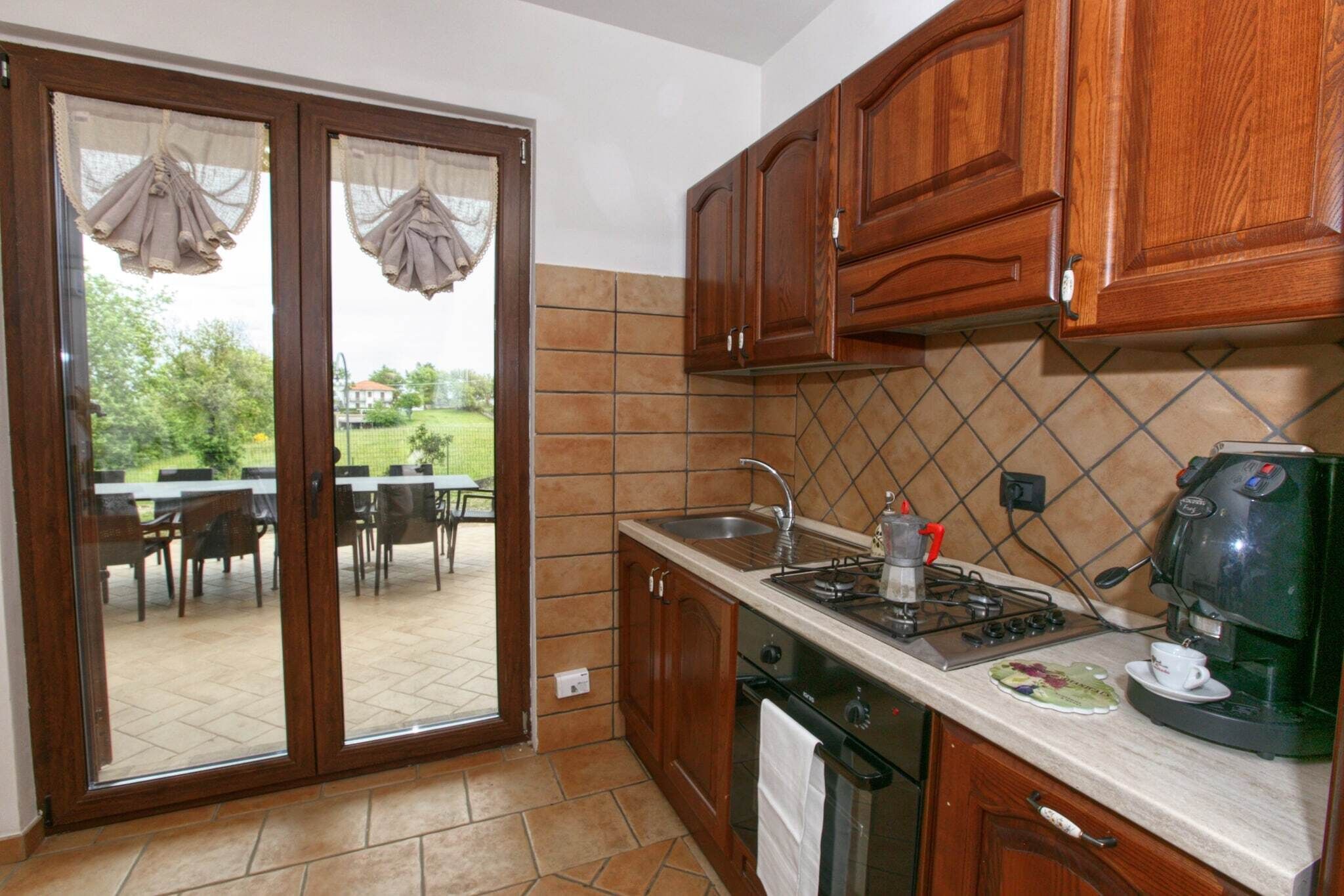 Cozinha com armários de madeira, fogão a gás, cafeteira e vista para um pátio externo com mesa e cadeiras.
